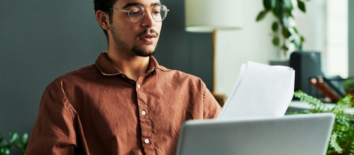 Young confident man with papers sitting in front of laptop